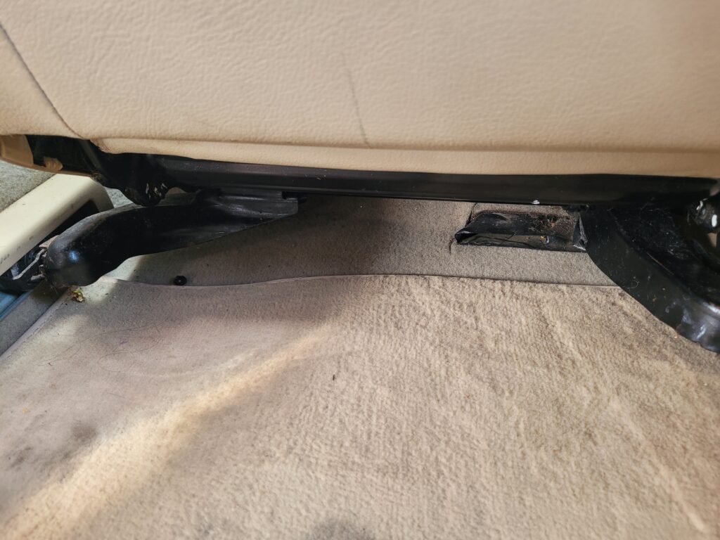 Picture of the underside of the driver's seat of a Volkswagen Jetta. Light tan coloured leather seat shown and light tan carpet with black, metal seat fixings in the centre of the image. 