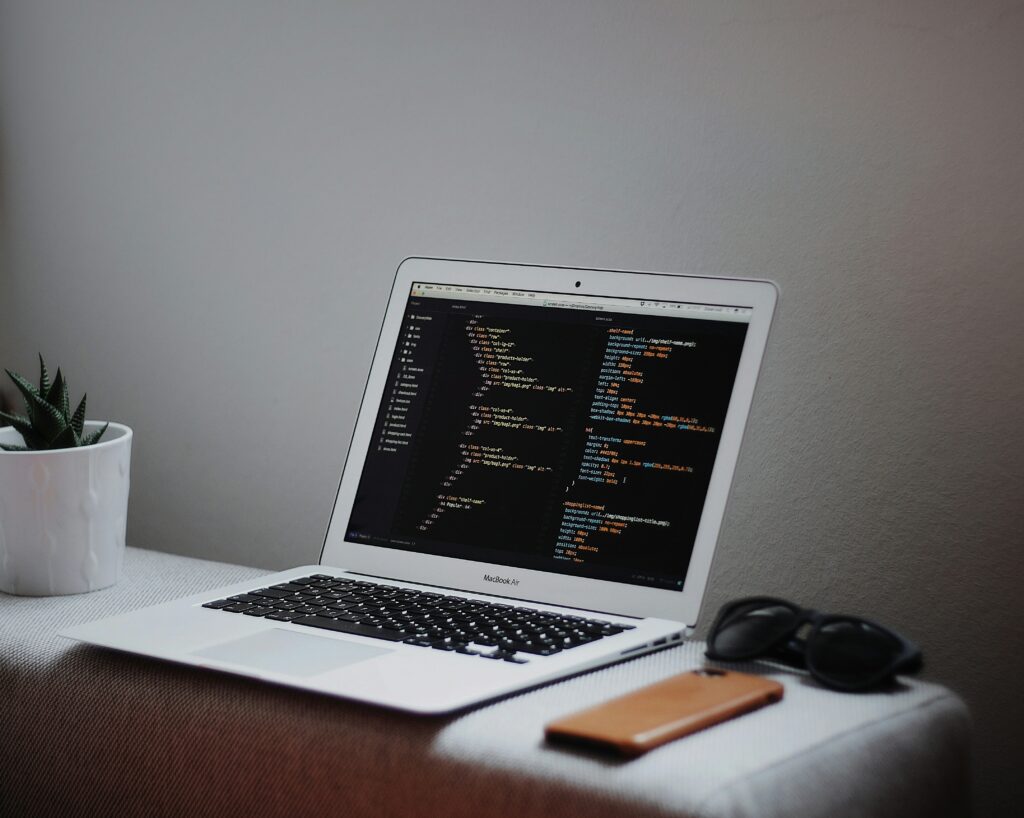 a picture of a silver Macbook pro sitting on a light grey ottoman open showing a screen that has light green, teal, and orange text running a coding sequence along the screen. On the left hand side of the picture is a small, dark green cactus-type plant in a small, white, textured pot. On the right-hand side of the laptop is a pair of black sunglasses and a phone in a tan, slim case facing down. 