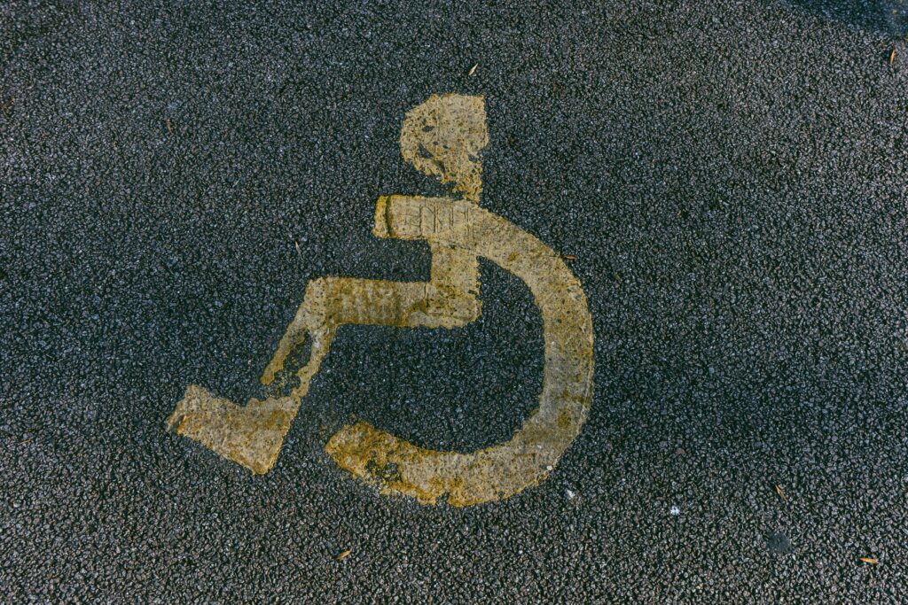 An image of a distressed yellow-coloured disability sign for a parking space. Symbol is of a stick person in a wheel chair painted on dark, clean pavement with the person facing to the left. 