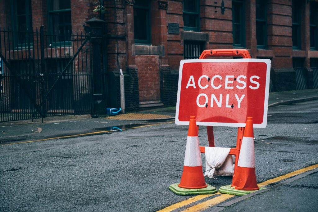 Foreground: A red, rectangular road sign that says "ACCESS ONLY" in white, capital letters with a white border around the edges. Two orange and reflective white traffic cones sit in front of the road sign in the middle of a two-lane city street. They sit partially on the double-into-single yellow line. 
Background: dark red brick building with a black metal parking lot fence on the side of the building. 