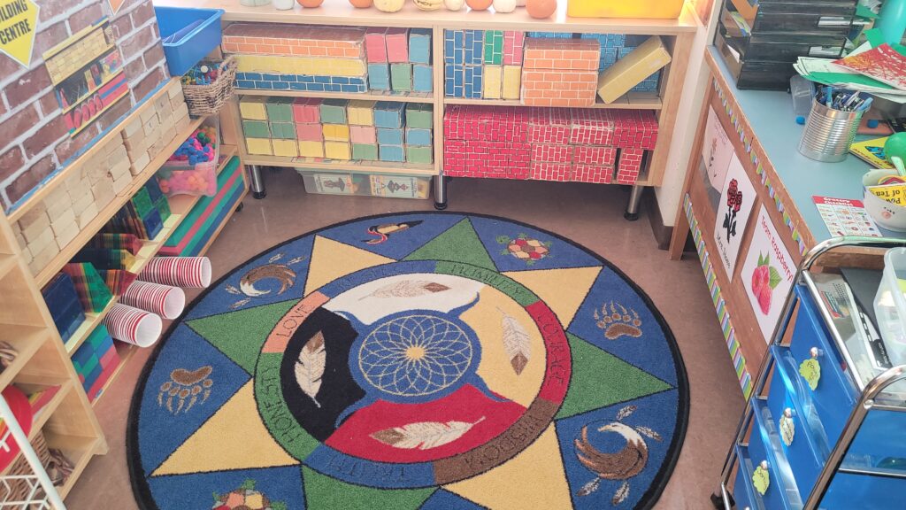 Colorful classroom corner with a circular rug featuring a Native American-inspired design. Shelves line the walls, filled with assorted learning materials and colorful books. Brightly decorated bulletin board on the right.