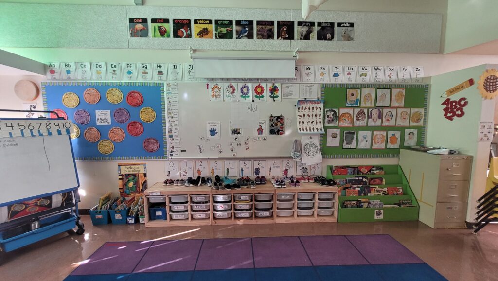 Colorful classroom corner with a rectangular rug featuring a Native American-inspired design. Shelves line the walls, filled with assorted learning materials and colorful books. Brightly decorated bulletin board on the right.