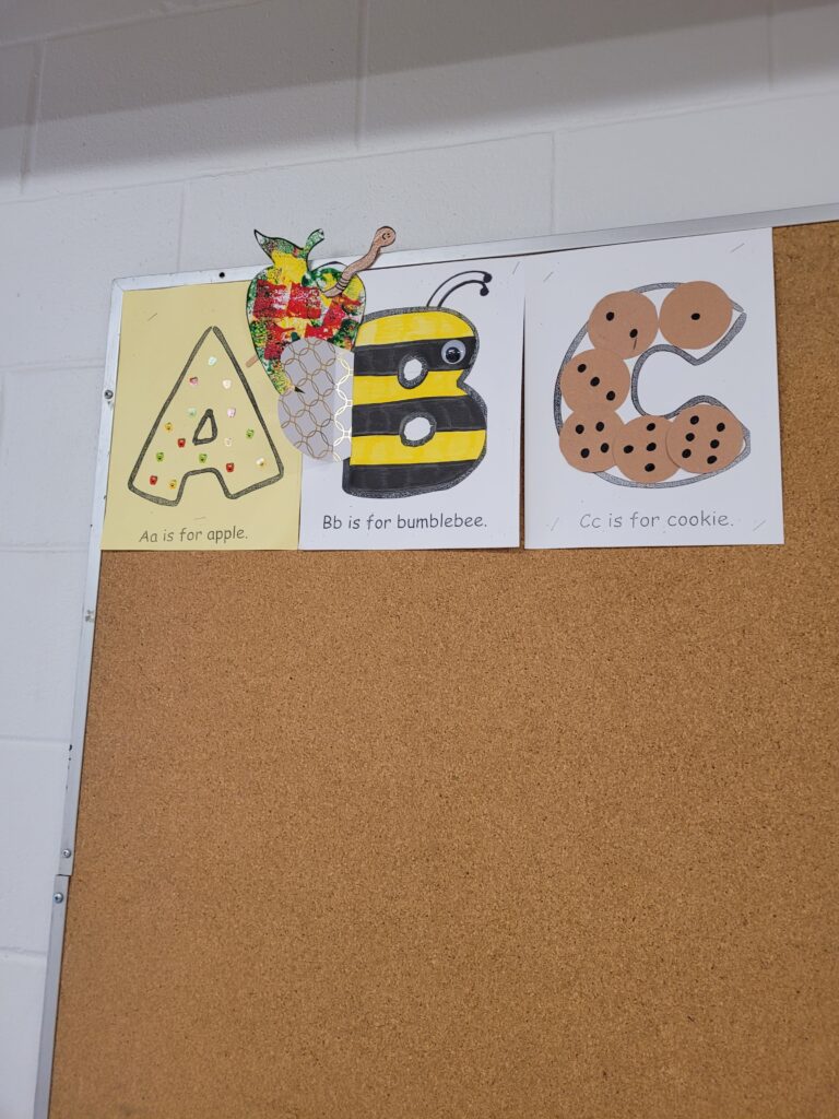 A classroom schedule on a whiteboard with illustrated magnets: carpet time, exercise time, math, snack, story time, music, lunch, quiet time, outside playtime, calendar time, centers, seat work, and recess. Alphabet posters are on the left.