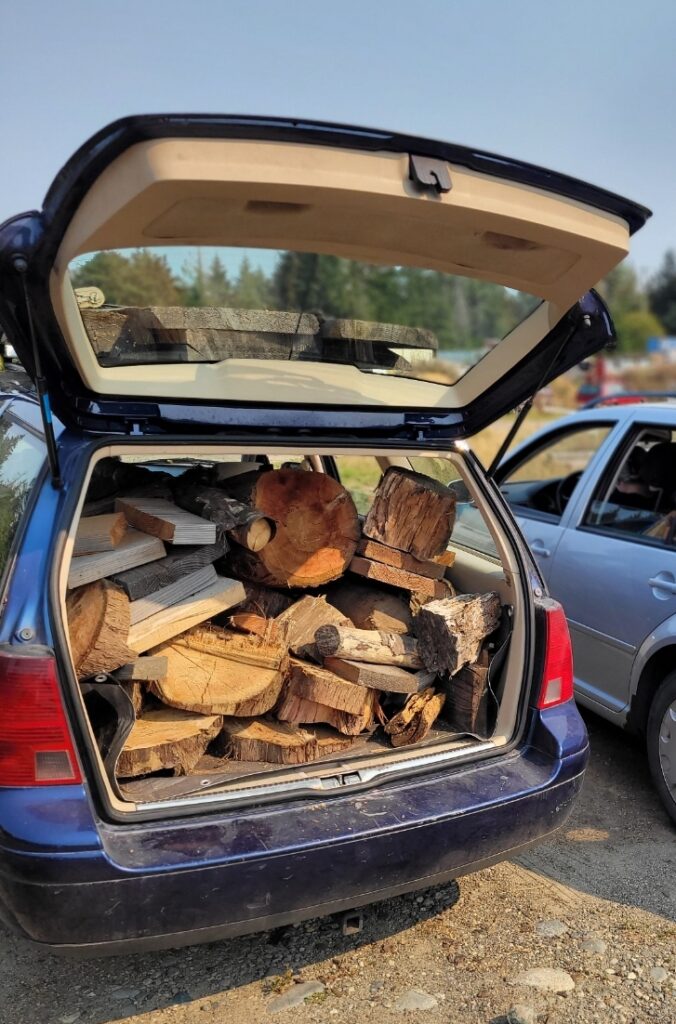 The image shows the back of a blue car with its trunk open, filled with large pieces of firewood. Another car is parked nearby, and trees can be seen in the background under a clear sky.