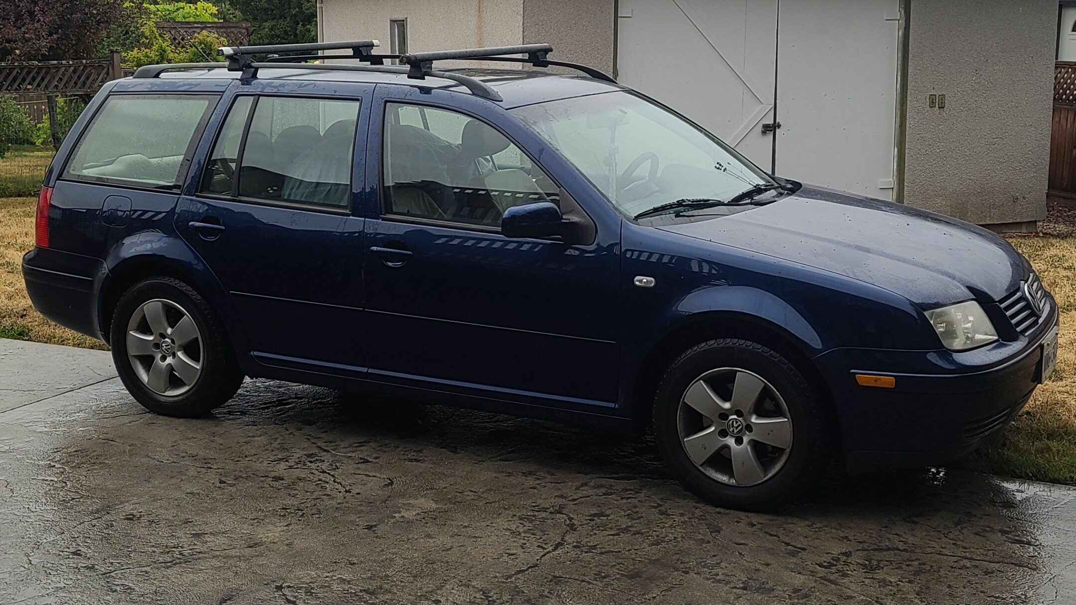 A dark blue station wagon is parked on a wet concrete driveway in front of a white garage. The car has roof racks and the surrounding area features some grass and a hedge.