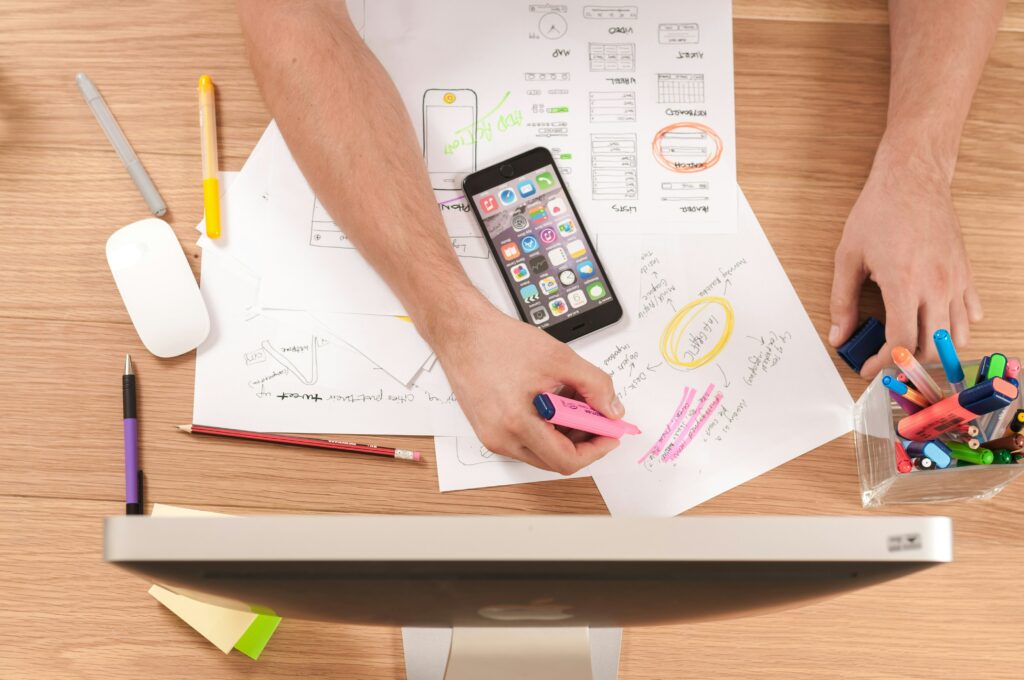 A person highlights notes on papers scattered on a desk. A smartphone displays app icons. Nearby, colorful pens and sticky notes are arranged. A computer monitor and mouse are visible, suggesting a workspace environment.