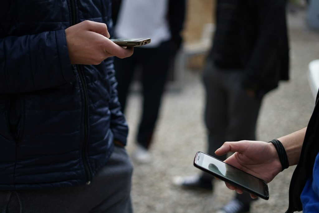 Two people are holding smartphones and standing outdoors. They are dressed in casual clothes, with one wearing a jacket and the other wearing a wristband. Other individuals are blurred in the background.