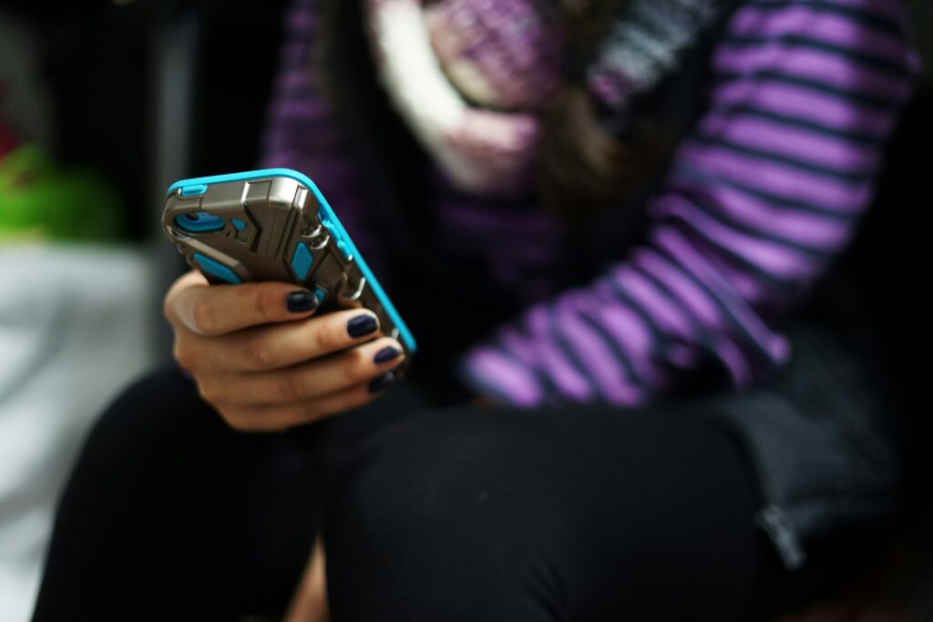 A person wearing a purple and black striped shirt and black pants is sitting and holding a smartphone with a blue and gray case. Their nails are painted dark, and a blurred background suggests an indoor setting.
      