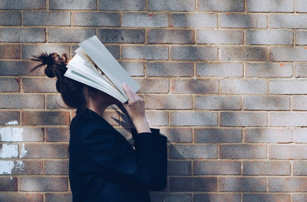 A person with long hair in a bun is standing against a brick wall, holding an open book in front of their face. They are wearing a dark jacket and appear to be immersed in reading. Light and shadow patterns are visible on the wall.