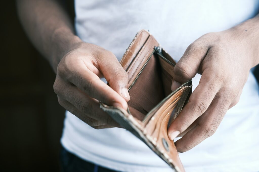 two rugged hands holding open a brown, worn leather wallet to reveal nothing inside the wallet, indicating a lack of money. The background shows the out-of-focus torso of a person that the hands belong to wearing a white short and black or dark blue jeans. 