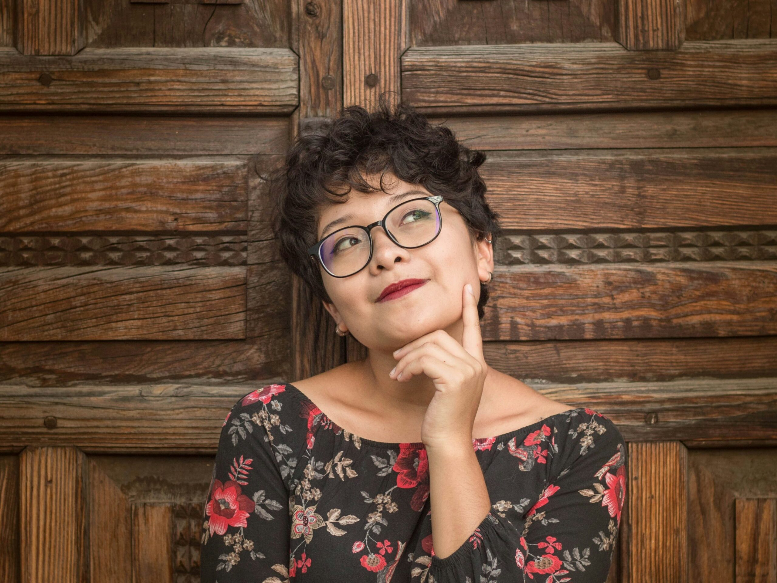 A woman leaning against a wood door, holding her chin and thinking