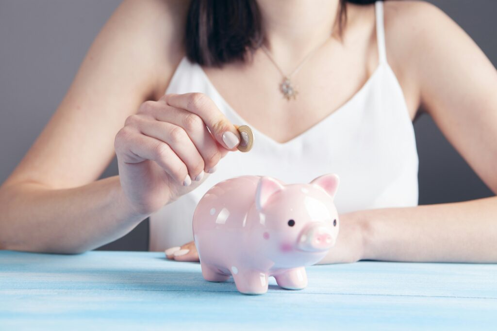 woman in white tank top placing a coin in a pink piggy bank on a blue table